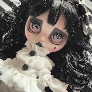Può includere: Una bambola con capelli neri, trucco scuro e un vestito bianco con bottoni neri. La bambola ha un incarnato pallido e un piccolo neo. Un fiocco a scacchi bianco e nero è tra i capelli della bambola. La bambola è su una superficie a strisce bianche e nere.