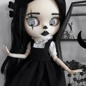 Può includere: Una bambola in stile gotico con capelli neri, trucco scuro e un vestito nero. La bambola ha grandi occhi con un design dettagliato. La bambola è posata davanti a una vetrina con oggetti decorativi. L'estetica generale è oscura e misteriosa.