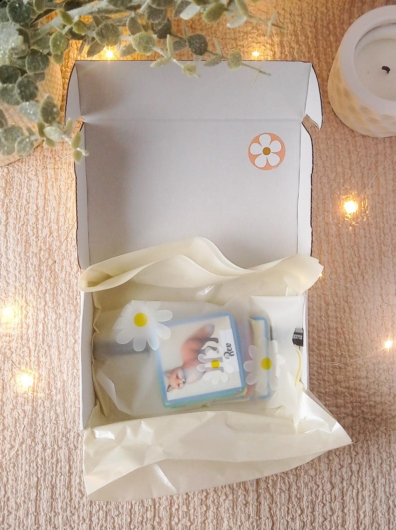 Puede incluir: Una caja de cart&oacute;n blanca con una pegatina de margarita blanca en la parte superior. La caja est&aacute; abierta y contiene una pila de tarjetas de colores con una ilustraci&oacute;n de zorro y la palabra "Rev" en una de las tarjetas. Las tarjetas est&aacute;n envueltas en papel de seda blanco.