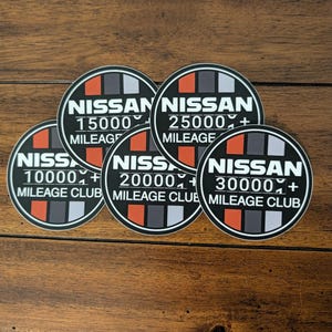 May include: Five circular stickers featuring the Nissan Mileage Club logo in black, white, orange, and gray. Each sticker displays the Nissan brand name, a specific mileage milestone of 10000, 15000, 20000, 25000, or 30000+, and the text Mileage Club.