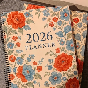 Op de afbeelding: Twee spiraalgebonden 2026-planners. De covers hebben een crèmekleurige achtergrond met een bloemenpatroon van oranje en blauwe bloemen en groene bladeren. De woorden "2026" en "PLANNER" zijn marineblauw gedrukt.