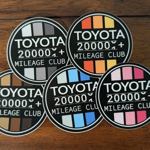 Puede incluir: Un conjunto de pegatinas redondas con el texto "TOYOTA 20000+ MILEAGE CLUB" en blanco sobre un fondo negro. Cada pegatina presenta un diseño de gradiente de color diferente, incluyendo tonos de gris, marrón, azul, naranja y rosa. Las pegatinas se muestran sobre una superficie de madera.