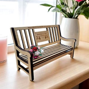 May include: A small wooden bench with a carved inscription that reads "Dedicated to the memory of Mum & Dad. Gone from our sight but not from our hearts." The bench is decorated with a small bouquet of pink and red flowers.