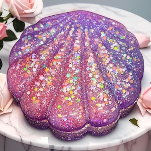 May include: A decorative, shell-shaped object with a gradient of pink and purple hues, heavily embellished with iridescent glitter. The object sits on a white marble surface, with pink roses in the background. The shell design is a unique decorative item.