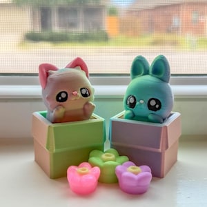May include: Two whimsical figurines: a cat with pink ears and a bunny with blue ears, each in a pastel box. Three flower-shaped decorations in pink, green, and purple are in front of the boxes. The figurines have large, expressive eyes.