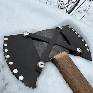 Puede incluir: Un hacha de doble filo negra con mango de madera y funda de cuero. El hacha está cubierta de nieve.