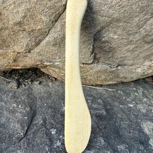 May include: A light-colored wooden butter spreader with a curved handle and a rounded end. The blade is wide and slightly curved. The spreader is made of natural wood and has visible grain.