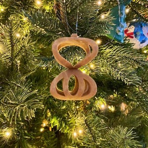 May include: A brown wooden Christmas ornament shaped like a figure eight with a decorative design. The ornament is hanging on a green Christmas tree.