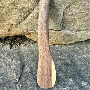 May include: A wooden butter spreader with a curved handle and a wide, rounded blade. The spreader is made of light brown wood with a visible grain pattern. The handle curves gently, and the blade is slightly angled.