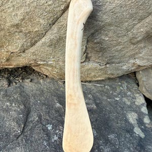 May include: A light-coloured, curved wooden spoon with a rounded handle and a wide, flat scoop. The spoon has a smooth finish and appears to be handmade. A small black marking is visible on the handle. The spoon is resting on a stone surface.