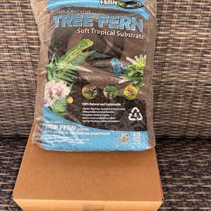 Peut inclure: Un sac en plastique transparent contenant du substrat tropical doux de fougère arborescente de Nouvelle-Zélande, posé sur une boîte en carton marron. Le sac présente des illustrations de reptiles et de plantes, et contient 10 litres.