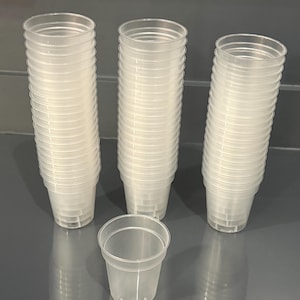 May include: A collection of clear, translucent plastic cups arranged in three tall vertical stacks with one additional single cup placed in the foreground. Each cup features a simple, cylindrical design with a smooth rim and a flat base.