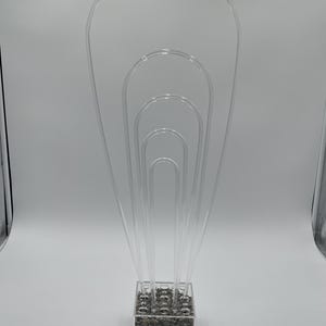 Könnte beinhalten: Transparenter Blumenarrangementshalter aus Acryl mit quadratischer Basis, gefüllt mit kleinen, dunkelgrauen Steinen. Das transparente Acryldesign weist mehrere bogenförmige Formen unterschiedlicher Größe auf.