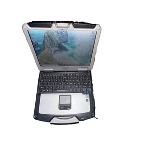 May include: A silver and black Panasonic Toughbook laptop with a black keyboard and a touchpad. The laptop is open and the screen is displaying a desktop background of a beach scene.