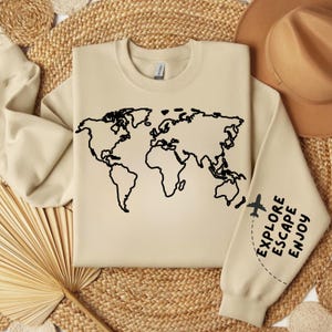 May include: A beige sweatshirt with a black outline of a world map and the text "Explore Escape Enjoy" on the sleeve. The sweatshirt is laid out on a woven surface.