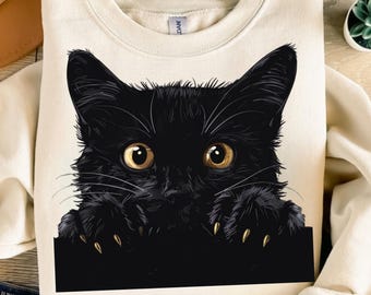 Op de afbeelding: Een zwarte kat met gele ogen kijkt over de rand van een crèmekleurig sweatshirt. De poten van de kat zijn zichtbaar en het sweatshirt heeft een ronde hals.