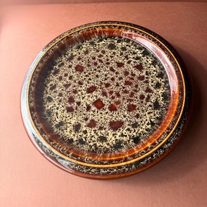 May include: A round ceramic plate featuring a speckled glaze pattern in shades of dark brown, black, and cream. The centre displays a dense, mottled texture that transitions into a smooth, solid dark brown rim along the outer edge of the plate.