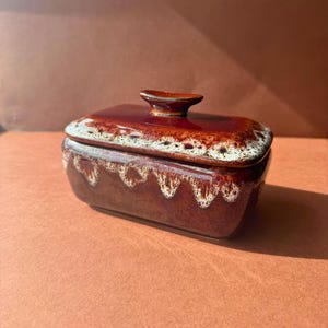 May include: A rectangular ceramic butter dish with a glossy dark brown glaze. The lid and base feature a decorative drip pattern in a creamy white colour along the edges. A small, oval-shaped handle sits centred on top of the lid for easy lifting.