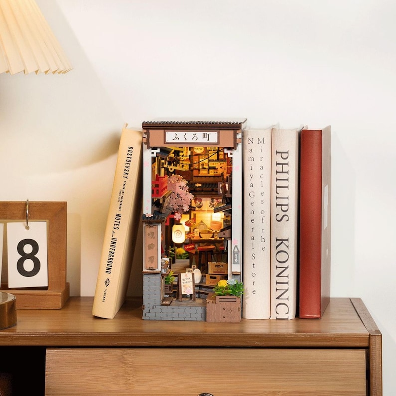 May include: A miniature Japanese street scene diorama, featuring a detailed facade with the text "ふくろ町". The diorama is illuminated and placed between books on a wooden surface, with a calendar showing the number 8.