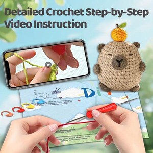 May include: A crochet tutorial image with a smartphone displaying a video. A crocheted bear with an orange on its head is next to the phone. The image also shows hands working with yarn and a crochet hook. Text at the top says "Detailed Crochet Step-by-Step Video Instruction."