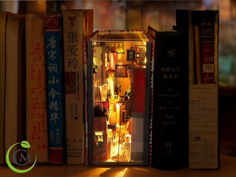 May include: A miniature dollhouse scene constructed inside a book. The dollhouse is illuminated from within and depicts a small alleyway with shops and buildings. The book is surrounded by other books on a shelf.