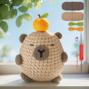 May include: A handmade crochet bear with an orange fruit on its head. The bear is light brown with darker brown accents and a dark brown nose. Crochet supplies, including yarn, a crochet hook, and stitch markers, are also visible.