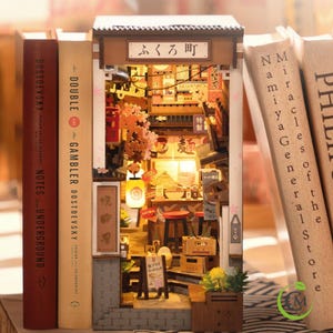 May include: A miniature Japanese street scene diorama, nestled between books. The detailed wooden structure features a restaurant with a glowing interior, cherry blossoms, and signage. The diorama is flanked by book spines with titles like "Notes from Underground" and "The Gambler."