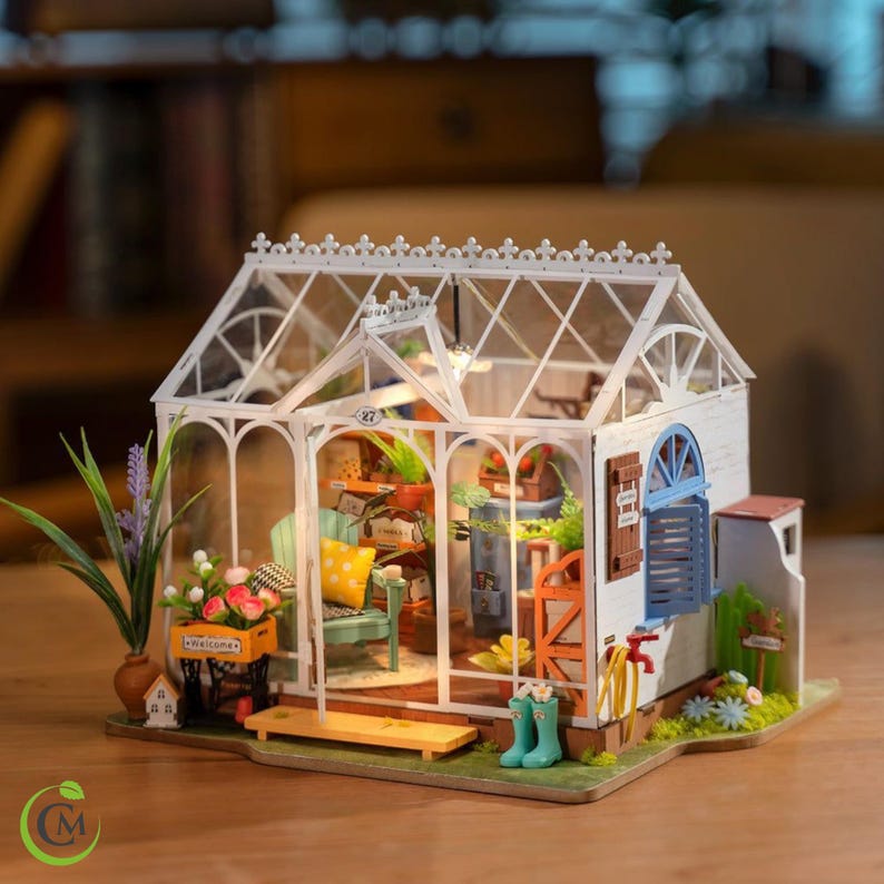 May include: A miniature greenhouse model with a white frame and clear roof. Inside, there are tiny plants, furniture, and decorative elements. The exterior features a small bench, blue boots, and a welcome sign. The overall design is detailed and charming.
