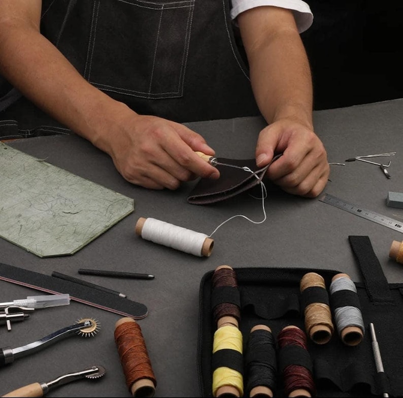 May include: A leatherworking kit with tools and supplies for stitching leather. The kit includes a variety of needles, thread, awls, and a ruler. The kit is laid out on a gray surface with a person stitching leather in the background.