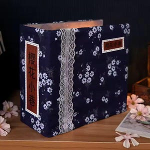 K&ouml;nnte beinhalten: Ein marineblaues Fotoalbum mit einem floralen Muster aus wei&szlig;en Bl&uuml;ten. Das Album hat eine wei&szlig;e Spitzenborte in der Mitte und rechteckige Etiketten mit japanischem Text. Das Album liegt auf einer Holzoberfl&auml;che.