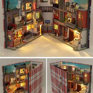 May include: A miniature model of a city street scene, built inside a book. The buildings are made of wood and painted in a variety of colours, including red, yellow, and blue. The street is lined with small trees and streetlights. The book is open and the model is illuminated from within.