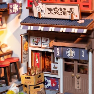 May include: A miniature wooden Japanese restaurant model with a blue awning and brown doors. The model features signs with Japanese text and illustrations of food and drink. There are also small wooden crates and red stools.
