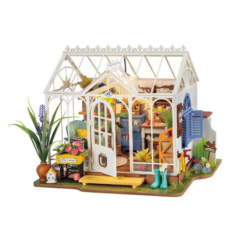 May include: A miniature greenhouse model with a white frame and glass panels. The interior features small furniture, plants, and decorative items. A sign reads "Welcome". The model includes a small house and a vase with lavender.