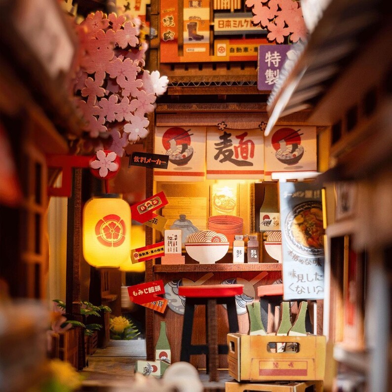 May include: Miniature Japanese ramen shop diorama with cherry blossom accents. The scene includes a red stool, a glowing lantern, and signs with Japanese text. A bowl of ramen sits on a wooden shelf, with bottles and a wooden crate.