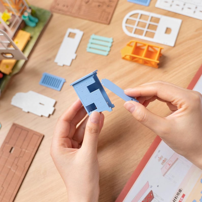 May include: A person assembling a miniature dollhouse. The image shows various unassembled parts in shades of blue, orange, white, and brown. The person is connecting a light blue cabinet piece to a matching blue component.