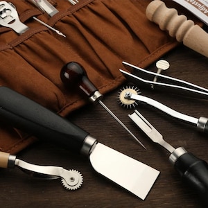 May include: A set of leather working tools, including a stitching awl, a stitching pony, a pricking iron, a stitching wheel, a leather skiver, and a leather punch. The tools are laid out on a brown leather roll.