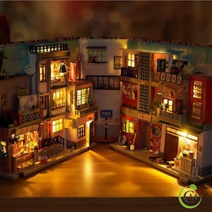 May include: A miniature dollhouse depicting a street scene, with several buildings featuring lit windows, balconies, and shop fronts. The buildings are painted in shades of red, brown, and yellow, with intricate details and signage. The dollhouse is illuminated with warm, yellow lights, creating a cosy and inviting atmosphere.