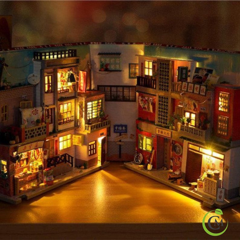 May include: A miniature dollhouse depicting a street scene, with several buildings featuring lit windows, balconies, and shop fronts. The buildings are painted in shades of red, brown, and yellow, with intricate details and signage. The dollhouse is illuminated with warm, yellow lights, creating a cosy and inviting atmosphere.