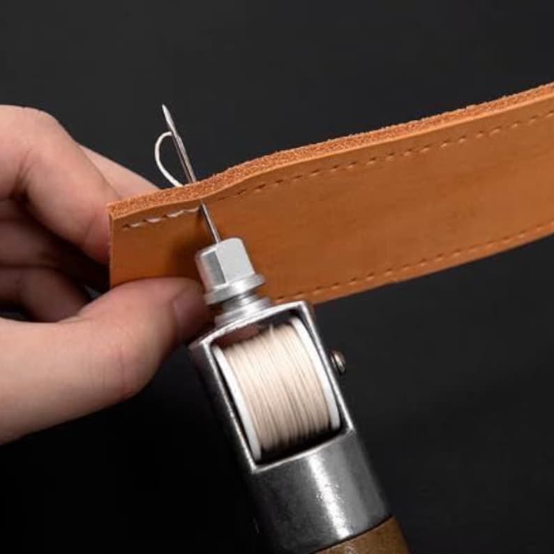Puede incluir: Primer plano de un proyecto de artesan&iacute;a en cuero en curso. Una herramienta de costura manual con una bobina de hilo blanco perfora un trozo de cuero marr&oacute;n claro. El cuero tiene agujeros de puntada pre-perforados.