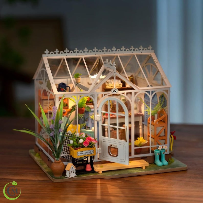 May include: A miniature greenhouse model with a white frame and glass panels. The interior is filled with tiny plants, flowers, and miniature gardening tools. A small sign reads "Welcome". The model includes a small wooden bench and a pair of blue boots.