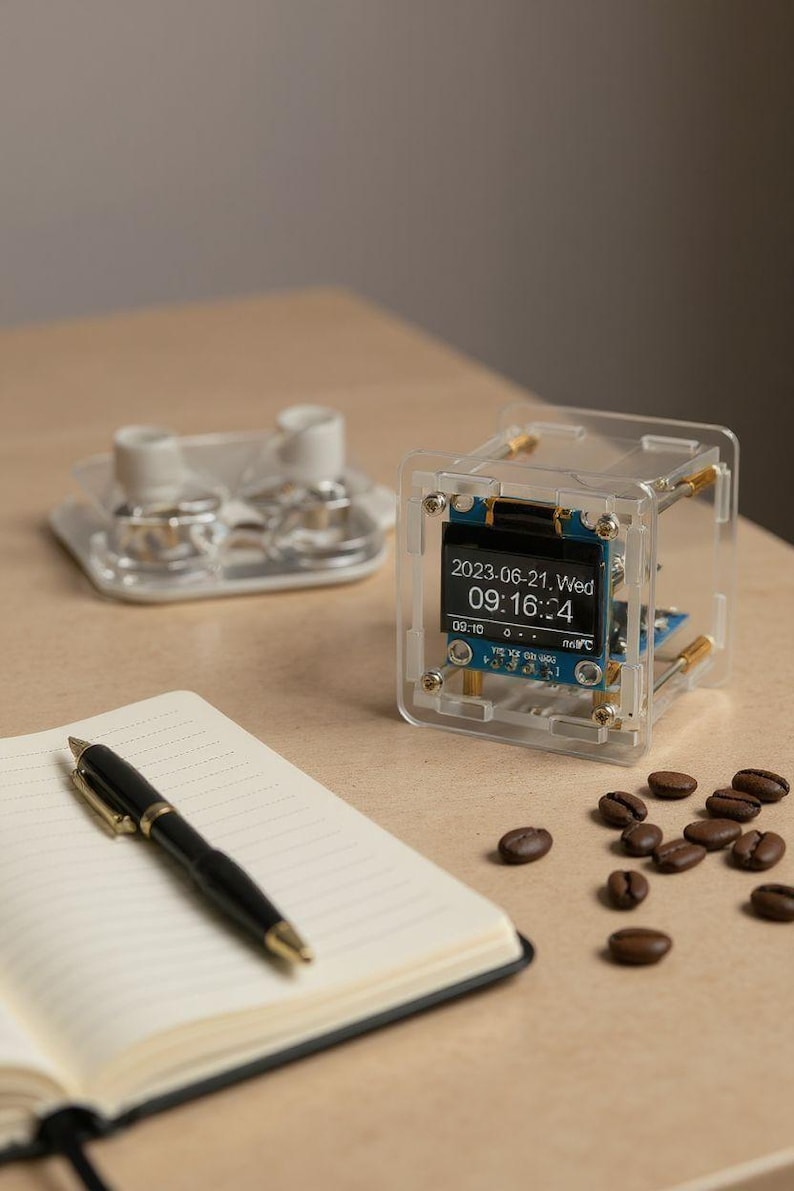 May include: A clear, cube-shaped electronic device with a digital display showing the date and time. A black pen rests on an open notebook. Coffee beans are scattered nearby. A clear tray with two small cups is in the background.