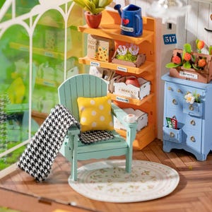 May include: Miniature dollhouse scene with a light blue Adirondack chair, a yellow polka dot pillow, and a black and white patterned blanket. A small orange shelf holds gardening supplies. A blue dresser with a drawer labeled "DAL" is also present.