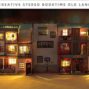 May include: A miniature model of a city street with three buildings, each with multiple windows and doors. The buildings are lit with warm white lights, creating a cosy and inviting atmosphere. The model is made of wood and paper and is detailed with small accents, such as signs and plants.