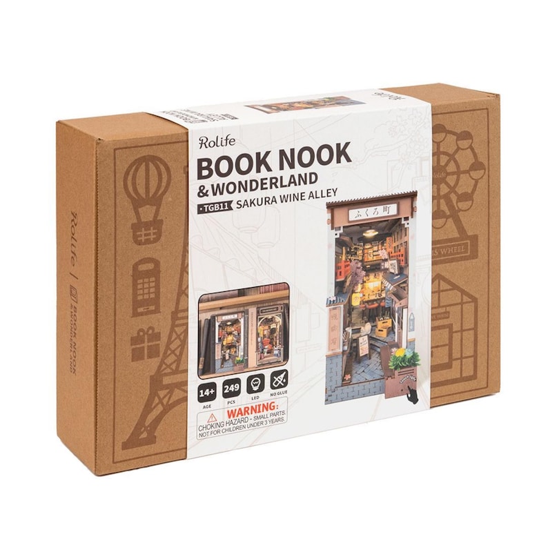 May include: A Rolife Book Nook & Wonderland kit, model TGB11, featuring a Sakura Wine Alley scene. The box is brown with illustrations of a hot air balloon, Eiffel Tower, and Ferris wheel. The kit includes 249 pieces and is for ages 14+.