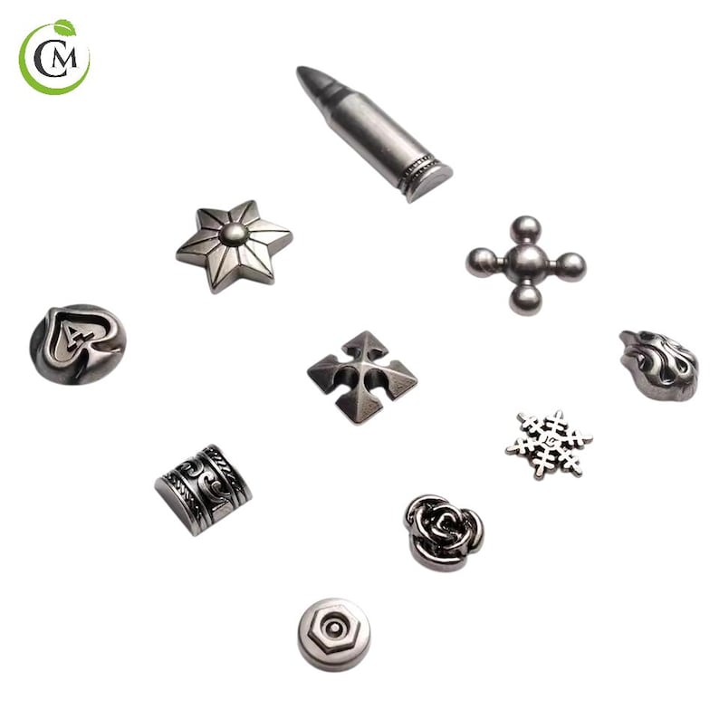 K&ouml;nnte beinhalten: Eine Sammlung silberfarbener Metallverzierungen in verschiedenen Formen, darunter eine Kugel, ein Stern, ein Kreuz und eine Rose. Die dekorativen Elemente sind auf wei&szlig;em Hintergrund angeordnet und zeigen ihre komplizierten Designs und Texturen.