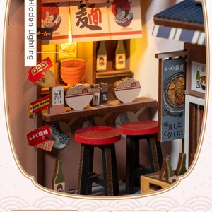 May include: Miniature Japanese ramen shop model with Hidden Lighting. The model features a counter with stools, bowls of ramen, and signs in Japanese. The shop has a warm, inviting glow from the interior lighting, creating a cozy atmosphere.