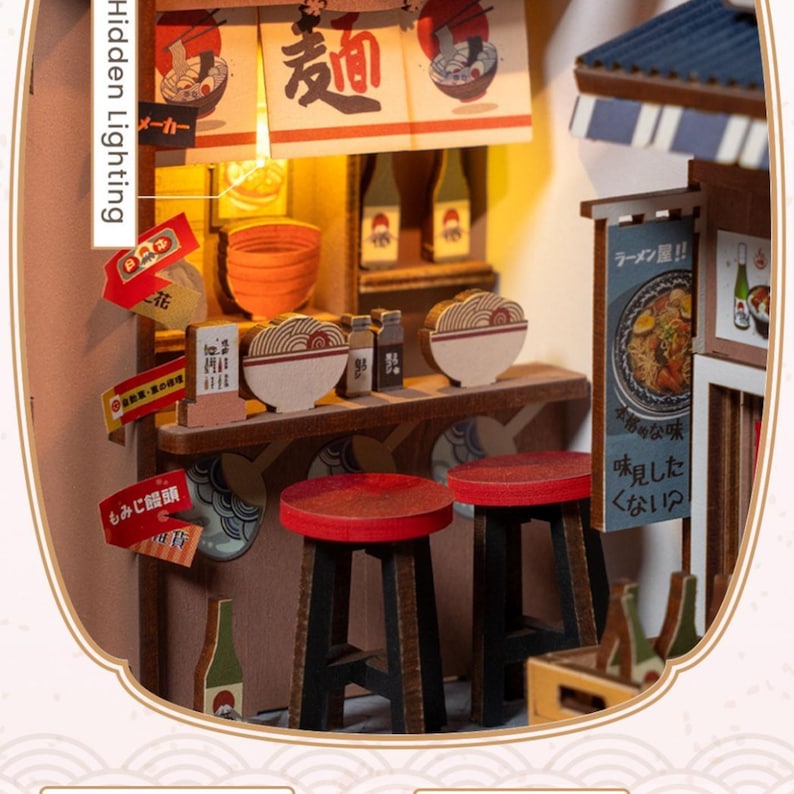 May include: Miniature Japanese ramen shop model with Hidden Lighting. The model features a counter with stools, bowls of ramen, and signs in Japanese. The shop has a warm, inviting glow from the interior lighting, creating a cozy atmosphere.
