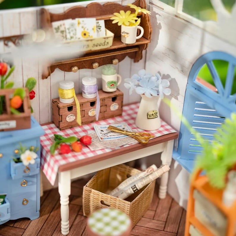 May include: Miniature dollhouse scene with a wooden shelf holding a mug with yellow flowers, and a small wooden box with spools of ribbon. A table with a red and white checkered tablecloth has a vase with blue flowers, scissors, and a basket underneath.