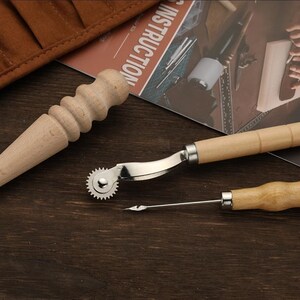 May include: A set of leather crafting tools including a stitching awl, a stitching groover, and a stitching wheel. The tools have wooden handles and are on a brown wooden surface.
