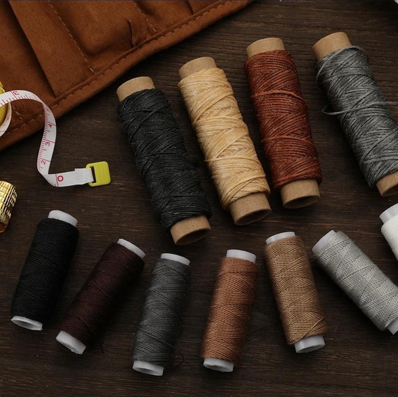 May include: Assortment of 12 spools of waxed thread in various colors, including black, brown, beige, gray, and white. The spools are arranged on a wooden surface.